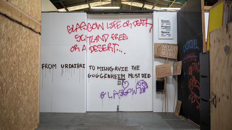 Installation view of the entrance to Govan Project Space, its white doors and walls spray-painted with protest-style graffiti reading "Glasgow Life or Death, Scotland Free or a Desert...", "From Urdaibai to Milngavie the Guggenheim must die" and "Glasgow", with handmade placards reading "Artists Against the Arsehole" and "All Hail Cultural Economic Development" propped against the wall beside a Govan Project Space sign.