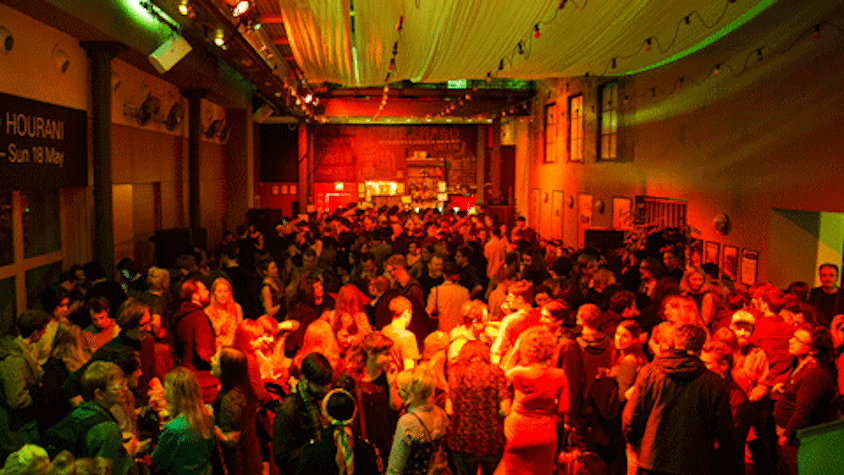 A large room with bright lights is packed with people with a bar at the end of the room. The board on top of the bar reads "Saramango: Cafe Bar". Long pieces of cloth are hanging across the ceiling, with a few rows of fairy lights.