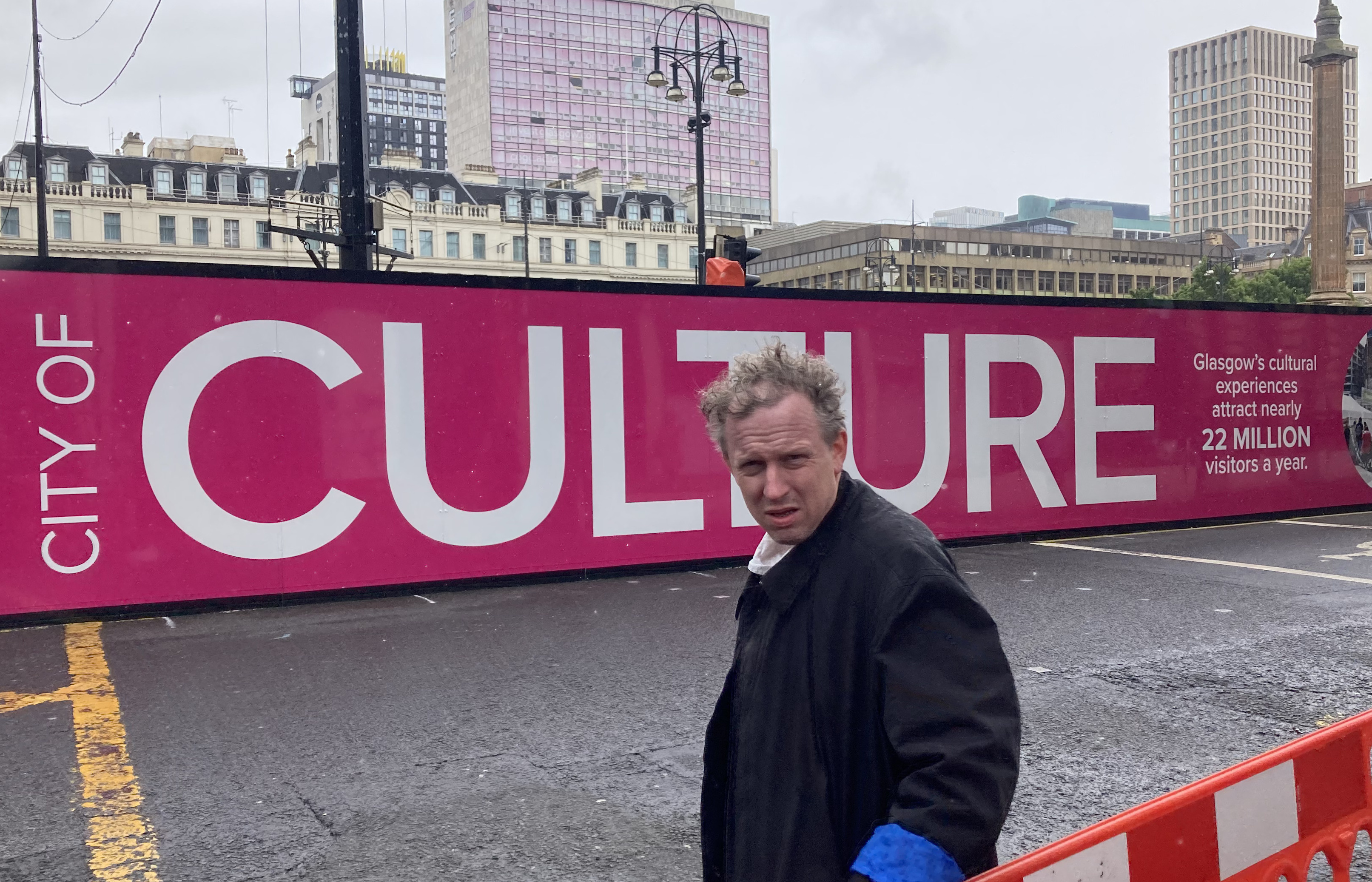 Colour photography of a man standing across from the George Square construction site, facing the camera. In the background, a pink banner reads "City of Culture." 