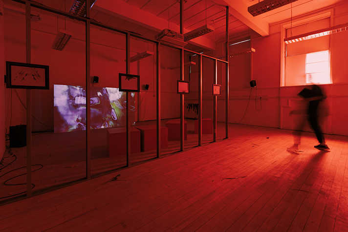 Film is projected onto a screen in a room lit in red. There is artwork displayed on wooden beams down the centre of the room.
