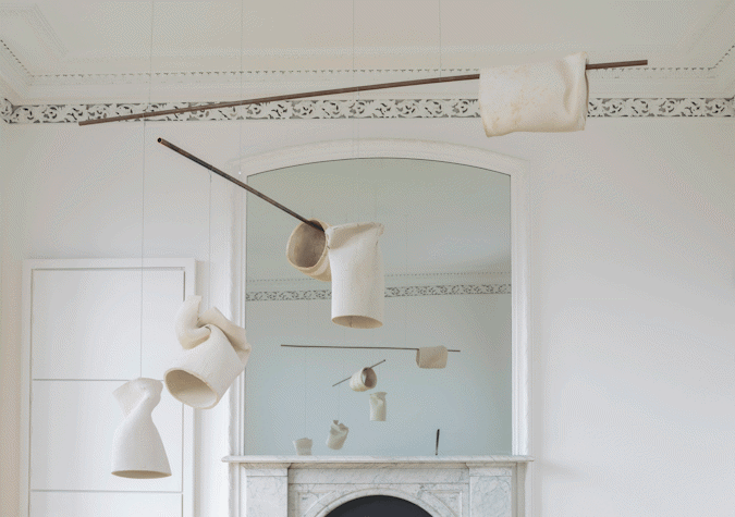 There is a white wall and a white fireplace with a mirror on top of it. There are ceramic copper tubes hanging from the mobiles in the ceiling.