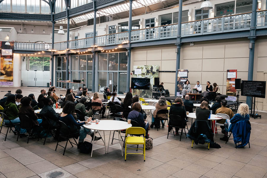 Wide view of an audience seated at round tables in a glass-roofed courtyard, facing a small panel discussion set up with a screen and banners in the background.
