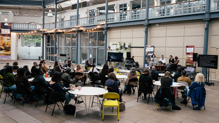 Wide view of an audience seated at round tables in a glass-roofed courtyard, facing a small panel discussion set up with a screen and banners in the background.