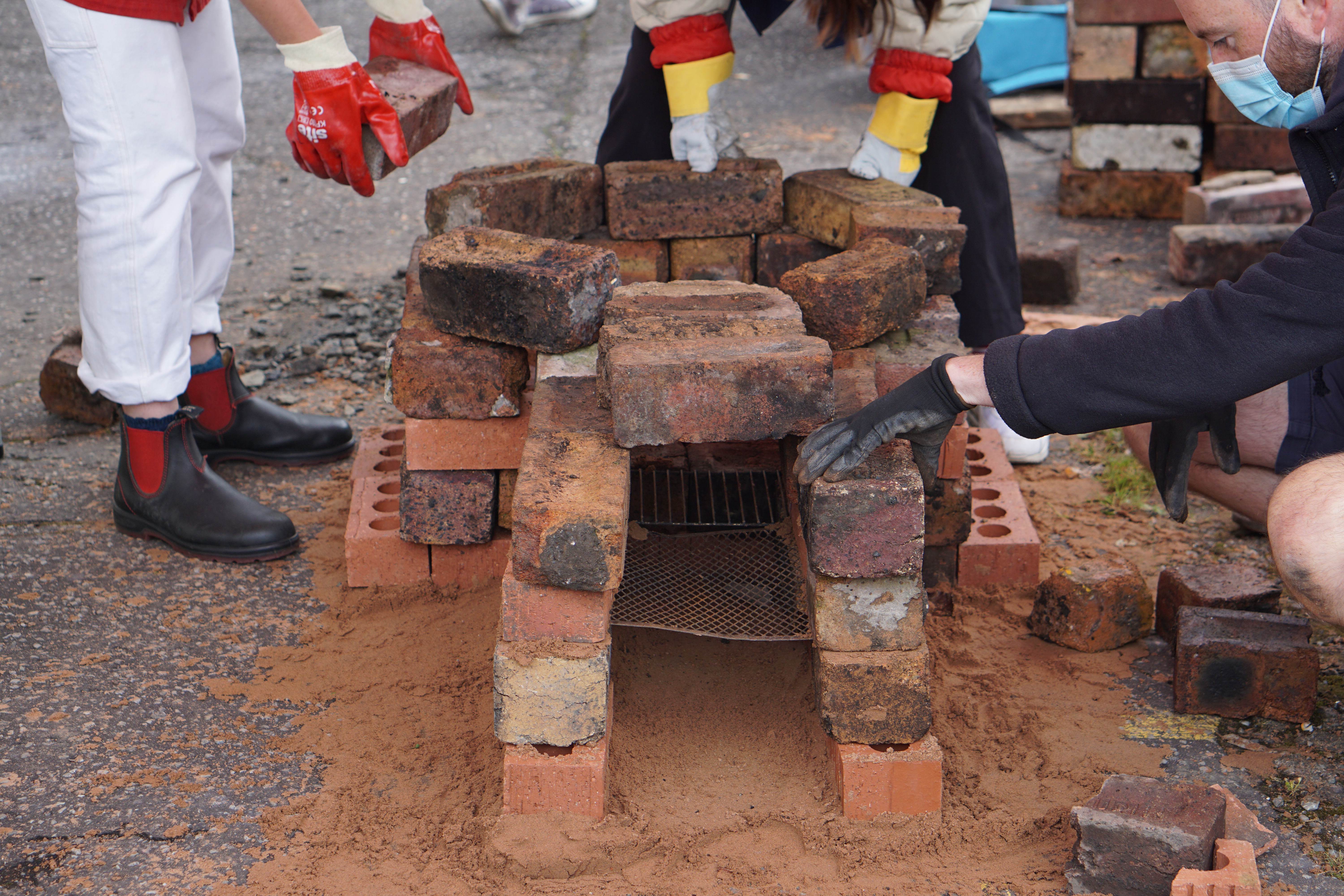 DIY Kiln Building. Close-up photography of three people's hands carrying bricks to add to a small outdoor brick kiln. The kiln sits on concrete with sand around its base, with metal trays and a circular brick section visible. 