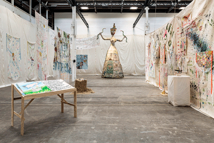Painted fabric is hung from walls in an exhibition space. At the back is a large sculpture of a figure.
