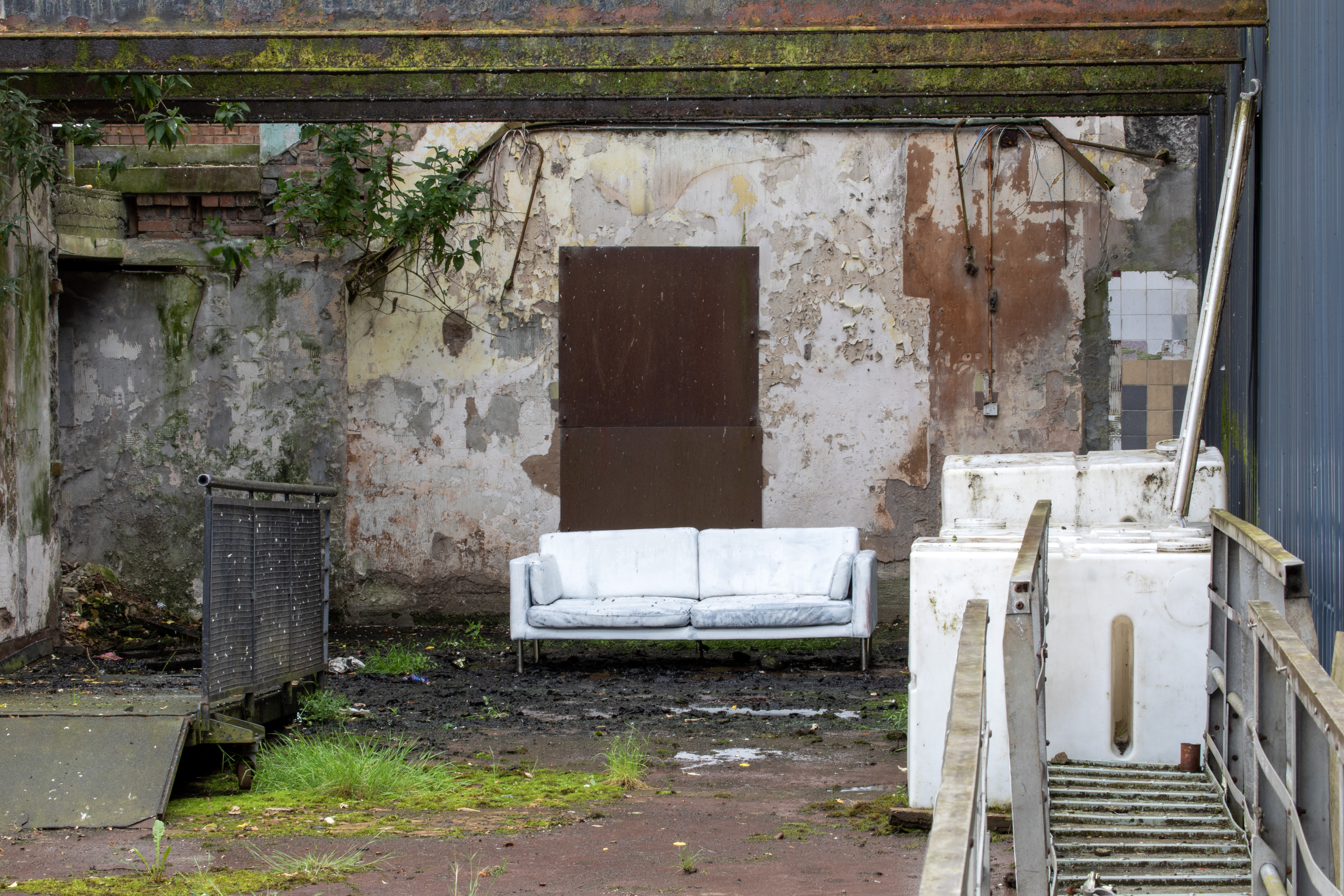 A weathered white sofa sits outdoors against a crumbling, mossy wall with a boarded-up doorway, beside discarded white appliances and a metal staircase.