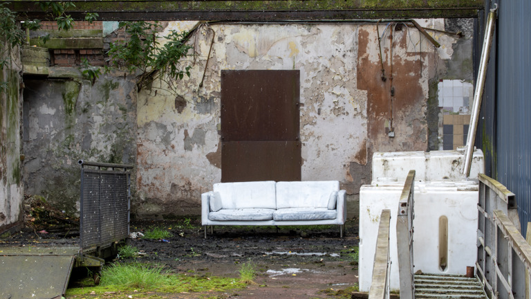 A weathered white sofa sits outdoors against a crumbling, mossy wall with a boarded-up doorway, beside discarded white appliances and a metal staircase.