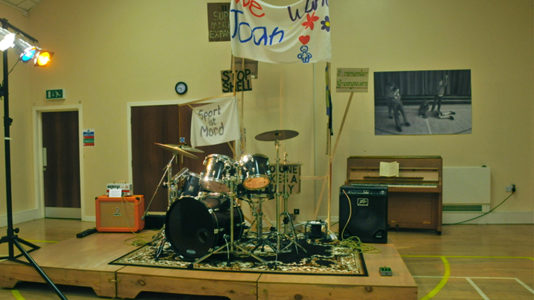 A drum kit in a village hall with a banner behind it 