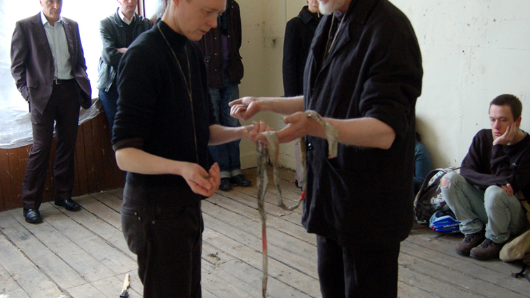 two people standing barefoot facing each other in room. they are holding a string of fabricant there are various wooden utensils on the floor surrounding the. people watch from the edge of the room.