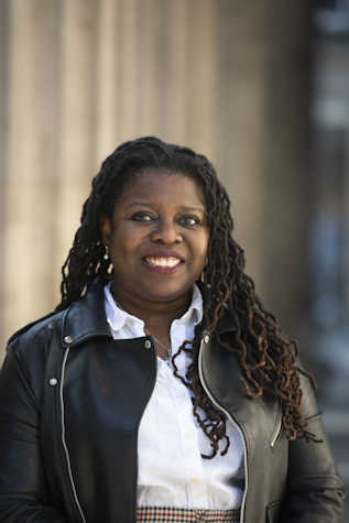 A woman with long black braids smiles, she is wearing a white shirt with a leather jacket over.