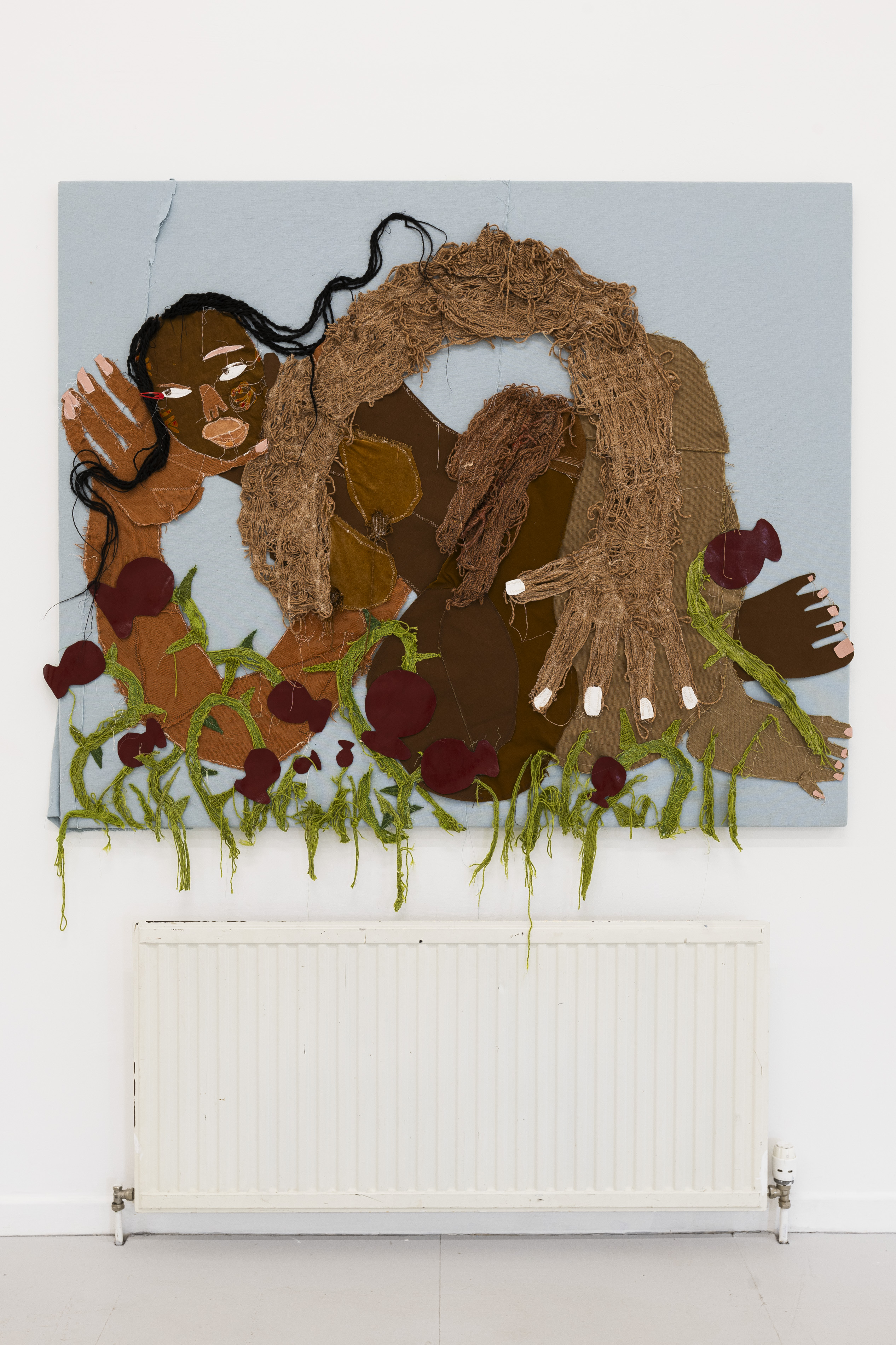 A mixed-media textile portrait of a reclining figure with long braided black hair and a mane of unravelled brown yarn, surrounded by dark red fruit-like appliqué shapes with trailing green stems, hung above a radiator.