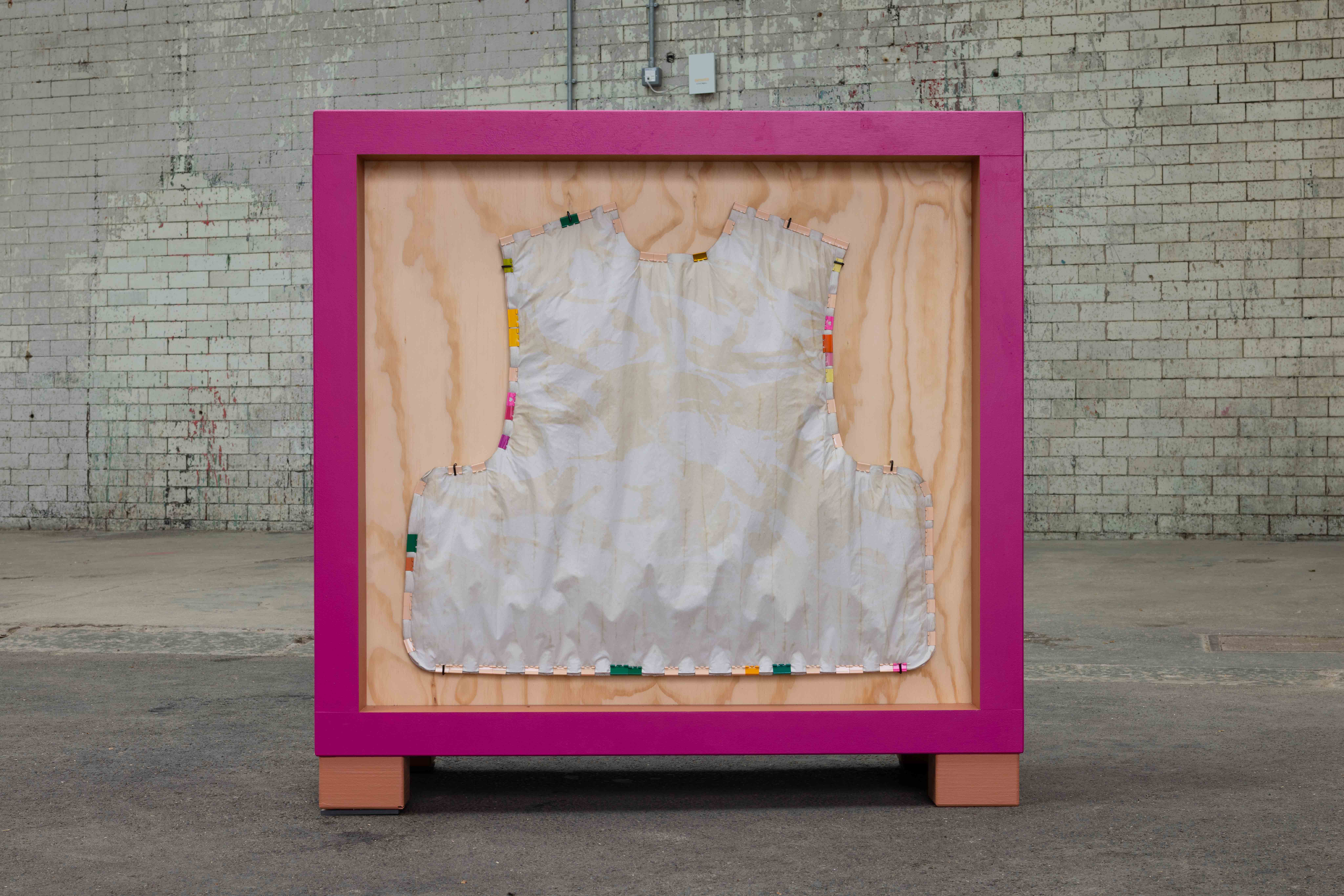 Magenta framed wooden crate sculpture displaying a single vest-shaped fabric form edged with colourful plastic clips, standing on a paved courtyard against a weathered brick wall.