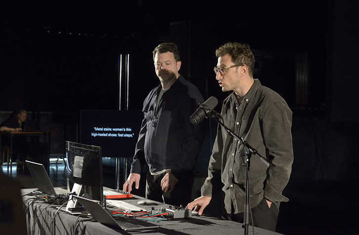 two people stood next to each other behind a desk. One is speaking into a microphone.