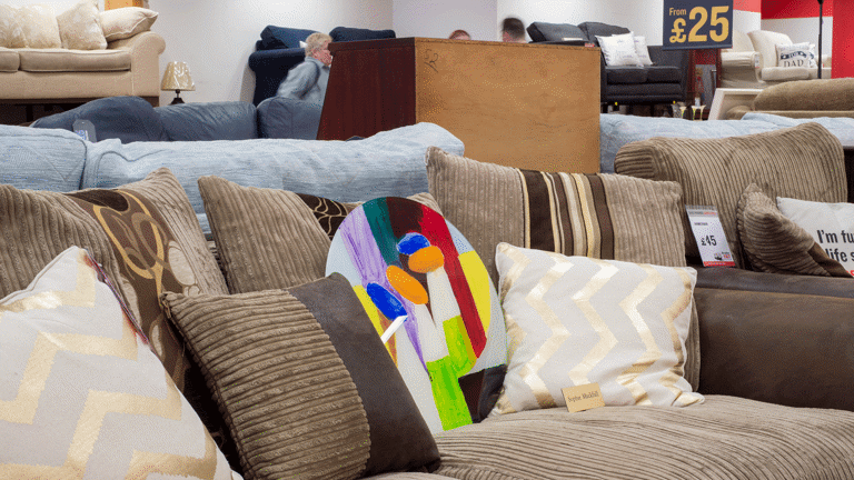 A colorful round painting is lodged in between a brown sofa with brown toned cushions in a British Heart Foundation charity shop.