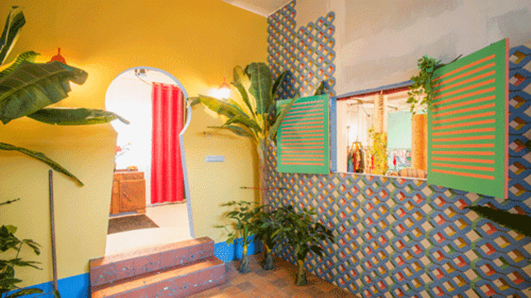 Installation view of a brightly coloured interior with plants, coloured walls and patterned wallpaper. There is a doorway in the shape of a keyhole, and a serving hatch 