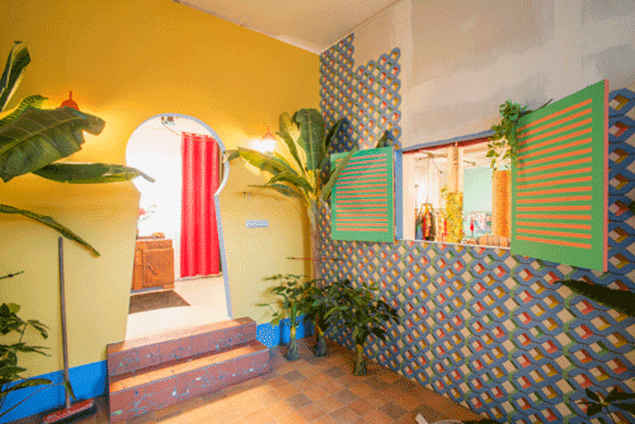 Installation view of a brightly coloured interior with plants, coloured walls and patterned wallpaper. There is a doorway in the shape of a keyhole, and a serving hatch