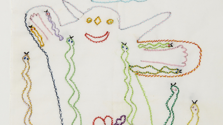 A colourful cross-stitch artwork on pale calico depicting the top half of a smiling creature. A cloud appears above while snakes of different size wind inside and outside the creature's outline. Two hears sit near the bottom, one pierced by an arrow.