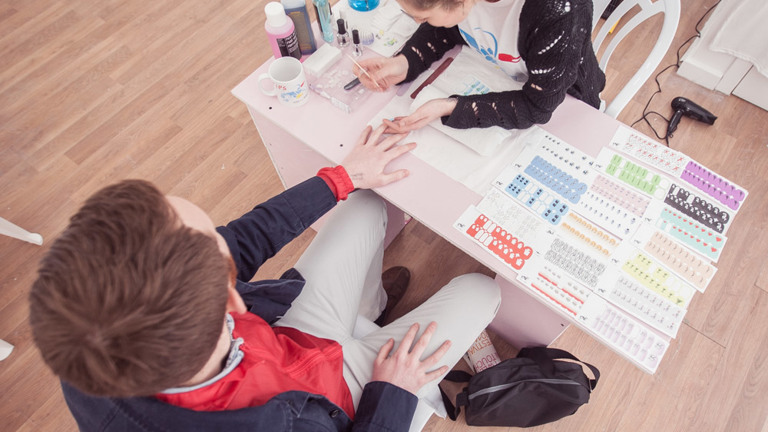 This is a top view of two people, a woman and a man, sitting at a small table. The table has multiple packs of nail stickers placed on the right side and nail kits on the left. These include three bottles and a sanitising cup, which holds a pair of scissors in it. The woman is holding a small metal stick in one hand and the man's hand in the other. 