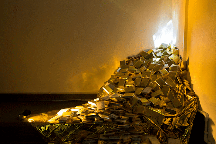 A pile of gold emergency blankets in their plastic packets sit in the corner of a dark room. A square of light shines at the top of the pile sending reflections out from the pile.