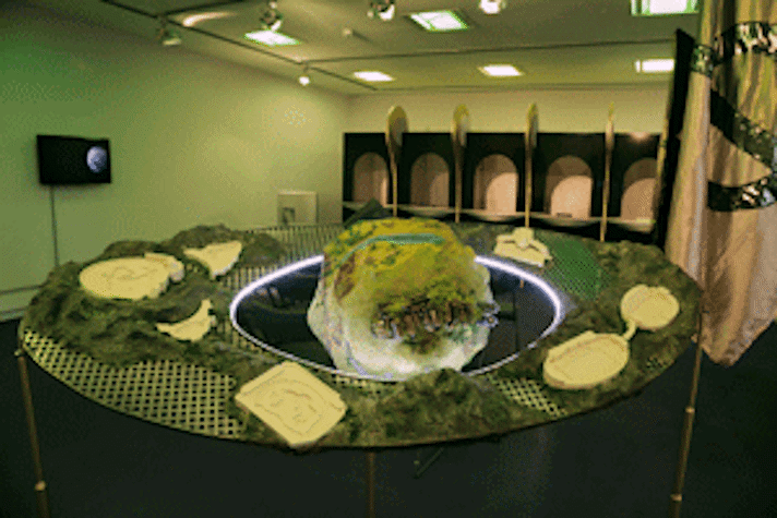 A large sculptural object in the foreground. A ring shaped table, with a lattice metal surface and a rough spherical object in the middle.