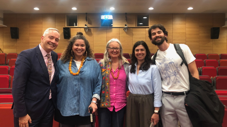 Five smiling people stand together in a lecture theatre with red seating, one using a walking stick, posing as a group after an event.