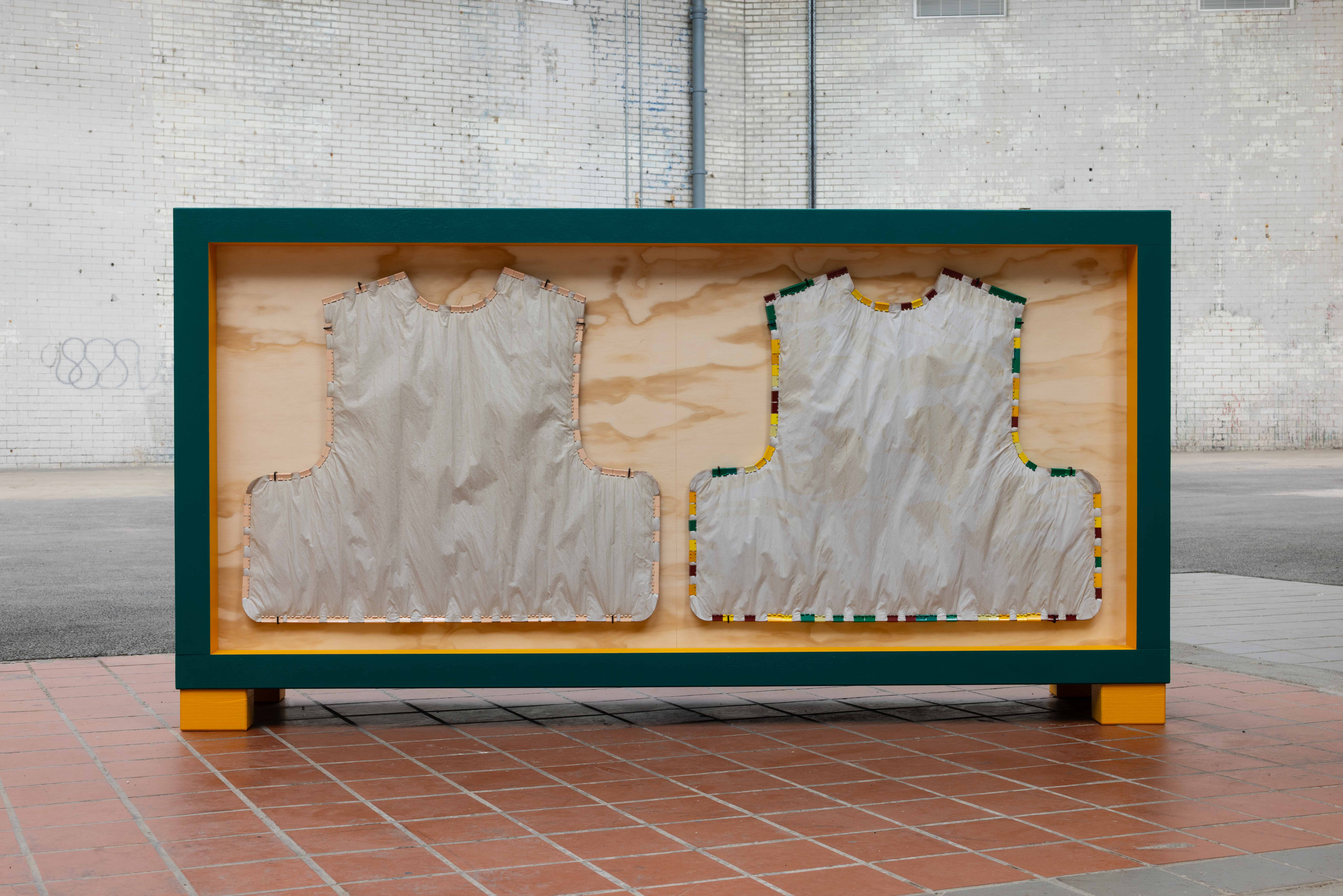 Green and orange framed wooden crate sculpture on red tiled ground, displaying two vest-shaped fabric forms edged with colourful plastic clips, set against a whitewashed brick wall.