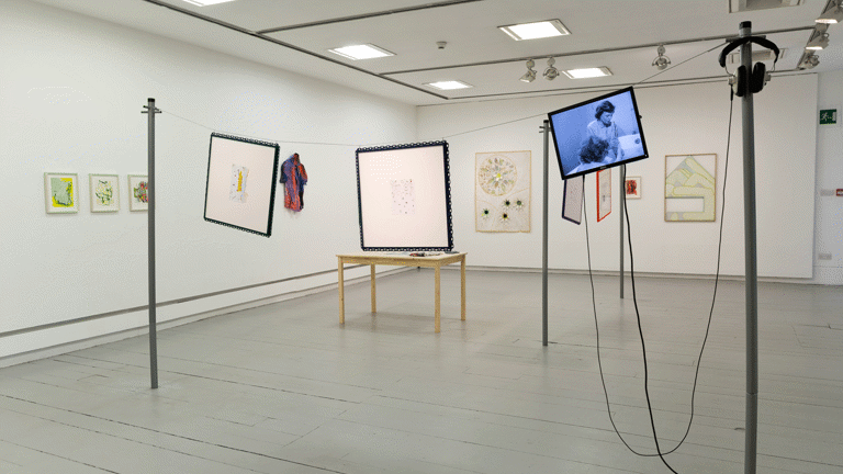 paintings hang on the walls of a gallery. in the middle of the space a wire hangs between two washing line poles, two artworks and a small tv monitor hang from the wire