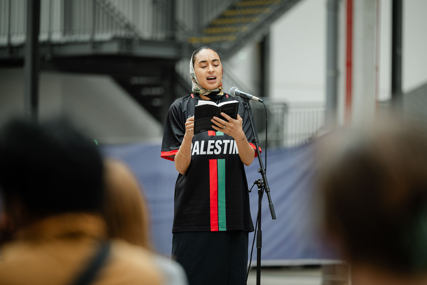 Close-up of a woman wearing a headscarf and a "Palestine" football shirt, standing and reading aloud from a book into a microphone, with a staircase and audience members visible in the background.