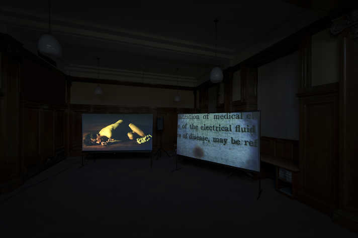 In a dark room two screens are forming a corner. The left screen has a white man lying on the floor in his underwear, in a fetal-like position. The right screen shows some text.