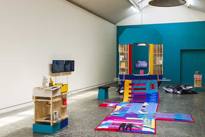 A well lit room with a large brightly coloured fabric artwork on the floor in the centre. On the left hand side are two wooden shelving structures covered in books, and two TV screens mounted to the wall. At the back of the room is a large wooden structure from which part of the fabric artwork is hanging.