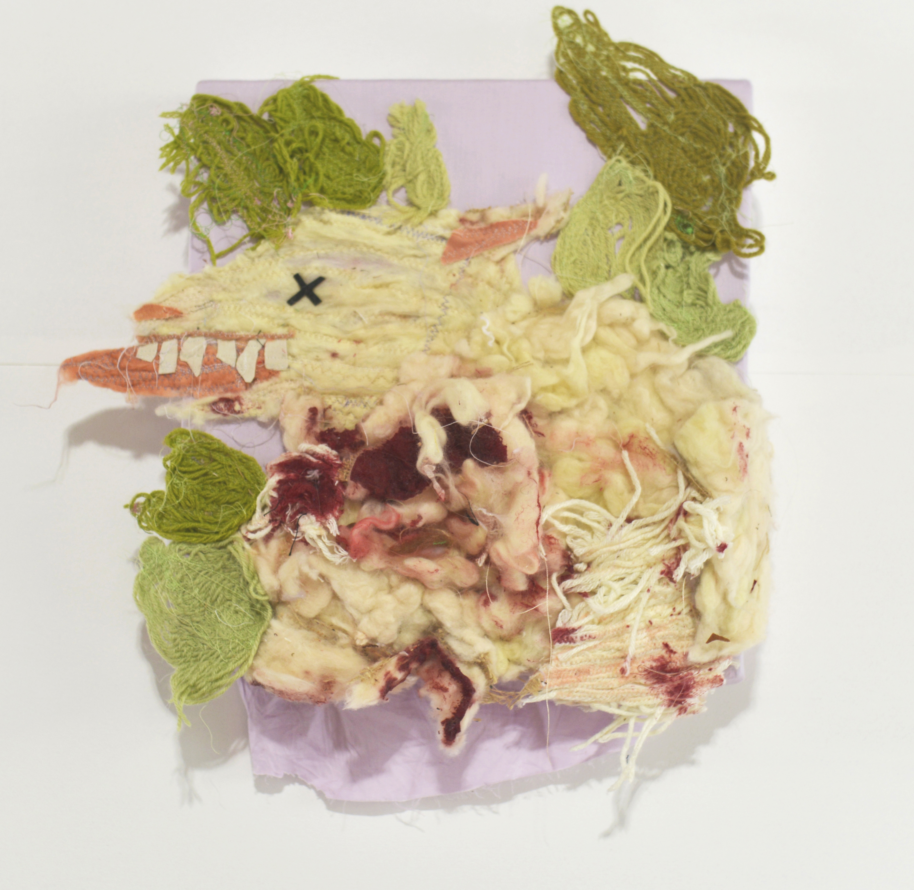 Textile work depicting the profile of a stylised dead sheep made from wool. Its tongue hands out and its eye forms a black cross. with red fabric suggesting blood along its neck. Green wooven leaves surround the sheep. 