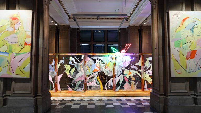 An abstract neon light showing two legs kicking the air in pointed heels, hangs at the top of a large wooden structure with glass panels and covered in fast gestural line paintings of figures and faces. The structure sits behind two stone columns on each of which a large scale canvas hangs, depicting fast gestural paintings of nude figures crouched and seated in pointed heels