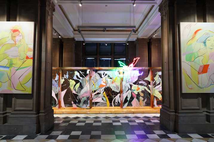 An abstract neon light showing two legs kicking the air in pointed heels, hangs at the top of a large wooden structure with glass panels and covered in fast gestural line paintings of figures and faces. The structure sits behind two stone columns on each of which a large scale canvas hangs, depicting fast gestural paintings of nude figures crouched and seated in pointed heels