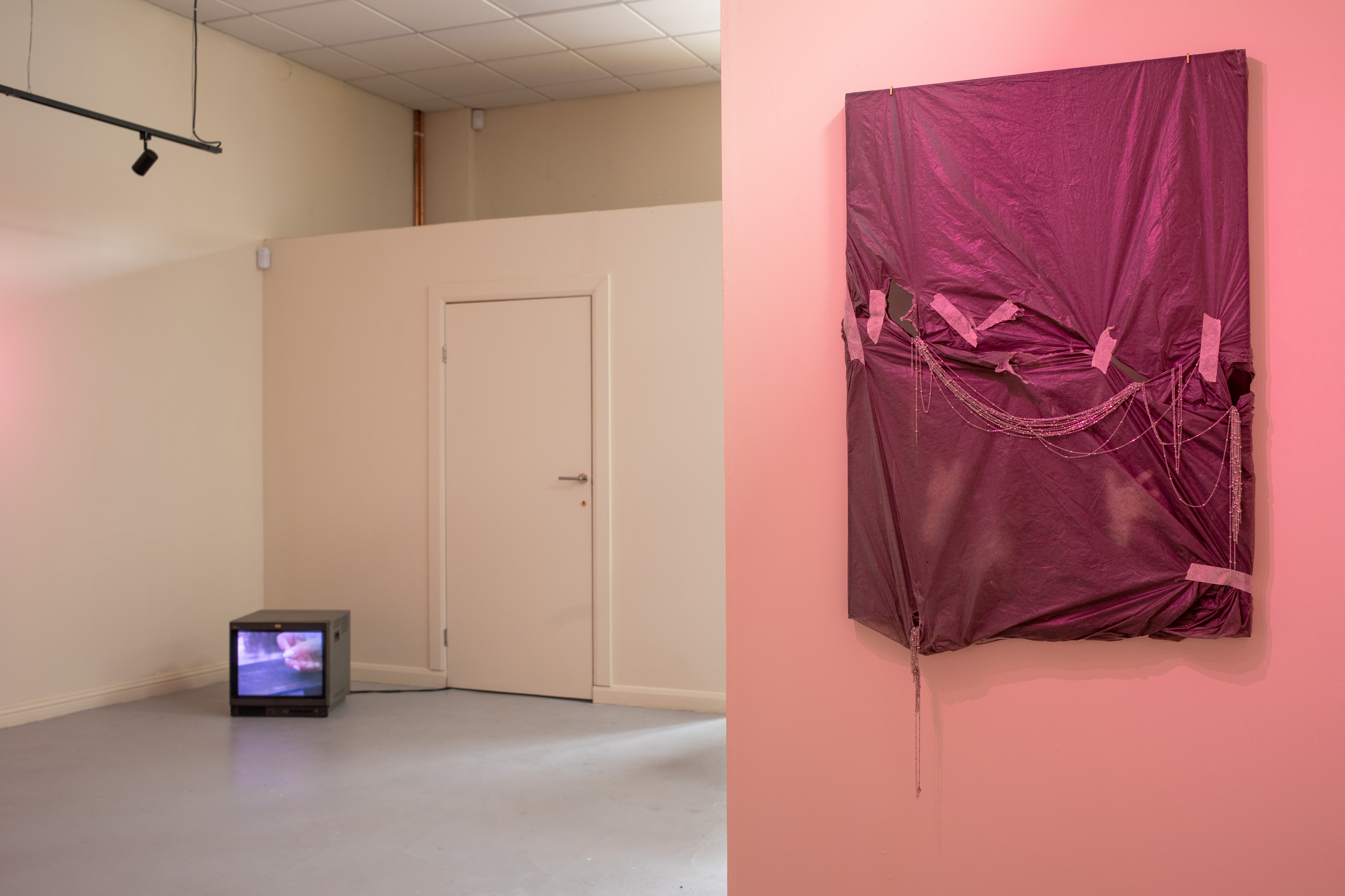 A gallery space at Gulabi Gallery showing a small CRT monitor playing video on the floor of a cream-walled room, alongside a draped, tape-patched magenta fabric work with beaded chains hanging from it on a pink wall.