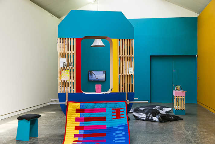 A well lit room with a fabric artwork hanging from a brightly coloured wooden structure.