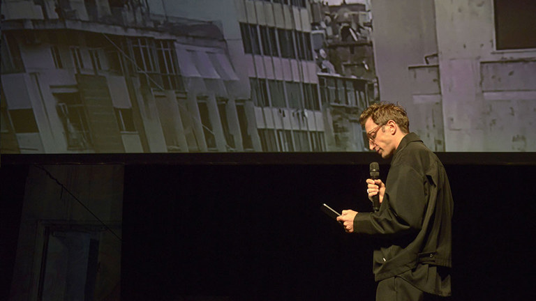 a figure with a microphone in front of a screen showing some buildings