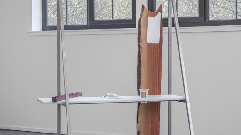 Sculptural work with metal frame, shelf and wooden plank in a gallery with a window behind