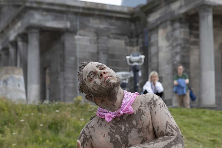 a close up of a person performing in front of a green hill with an old stone building on top. He is covered with a muddy mixture, wearing a pink neckerchief with a bare chest. His head is tilted, his eyes are closed and he looks blissful. There are two people watching him in the background. 