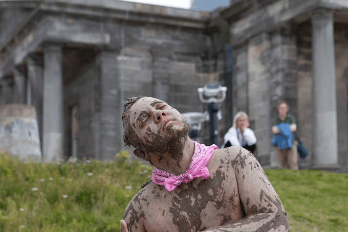 a close up of a person performing in front of a green hill with an old stone building on top. He is covered with a muddy mixture, wearing a pink neckerchief with a bare chest. His head is tilted, his eyes are closed and he looks blissful. There are two people watching him in the background.
