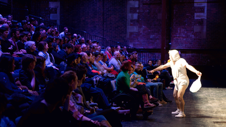 There is stage and an audience sitting in front of the stage. AA man wearing a baby diaper and clothes with a baby cap, has a whistle in his mouth and a plastic bag in one of his hands. His other hand is trying to reach out to a box that an audience member is holding. There are random audience members holding children's toys, including a toy telephone.