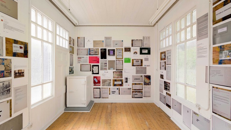 colourful photocopied sheets are pasted to the walls of a small gallery space. The sheets show letters, emails documents and other text-based media.