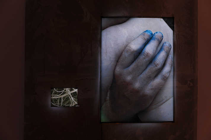 In a dark room, a large lit artwork featuring a hand with blue fingertips is next to a smaller artwork.
