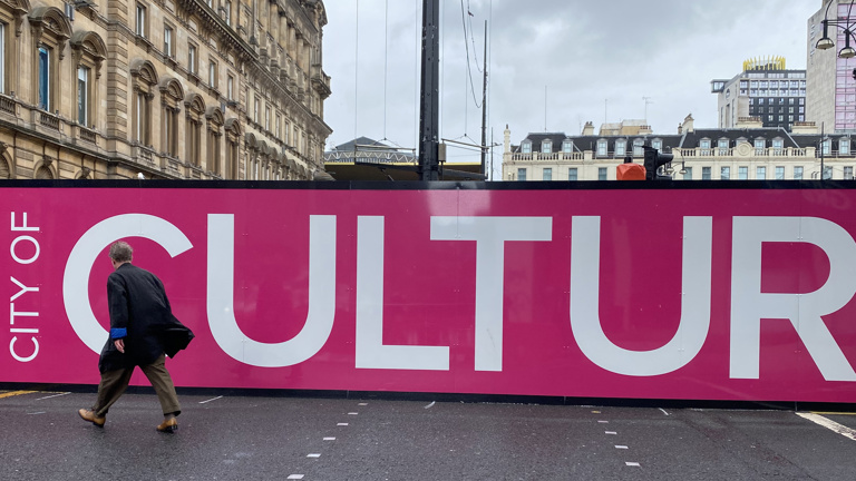 Colour photograph of a man walking away from the camera, towards the George Sqaure construction site. In the background, a pink banner reads "City of Culture." 