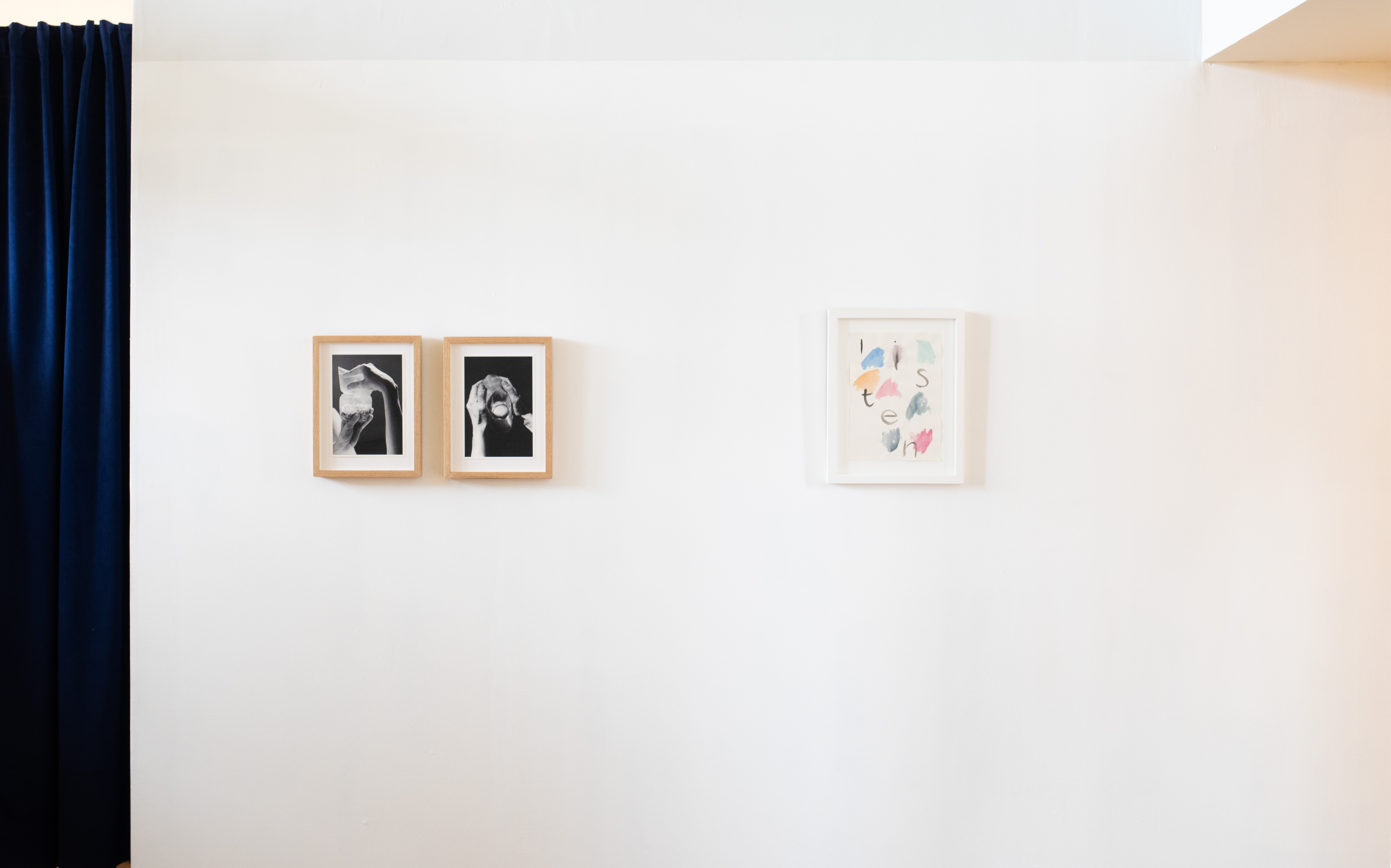 Two black-and-white photographs in oak frames show a hand holding a small round object close to the mouth. To the right, a separate white-framed piece spells out "listen" in loose cut-out letters interspersed with painted colour marks.