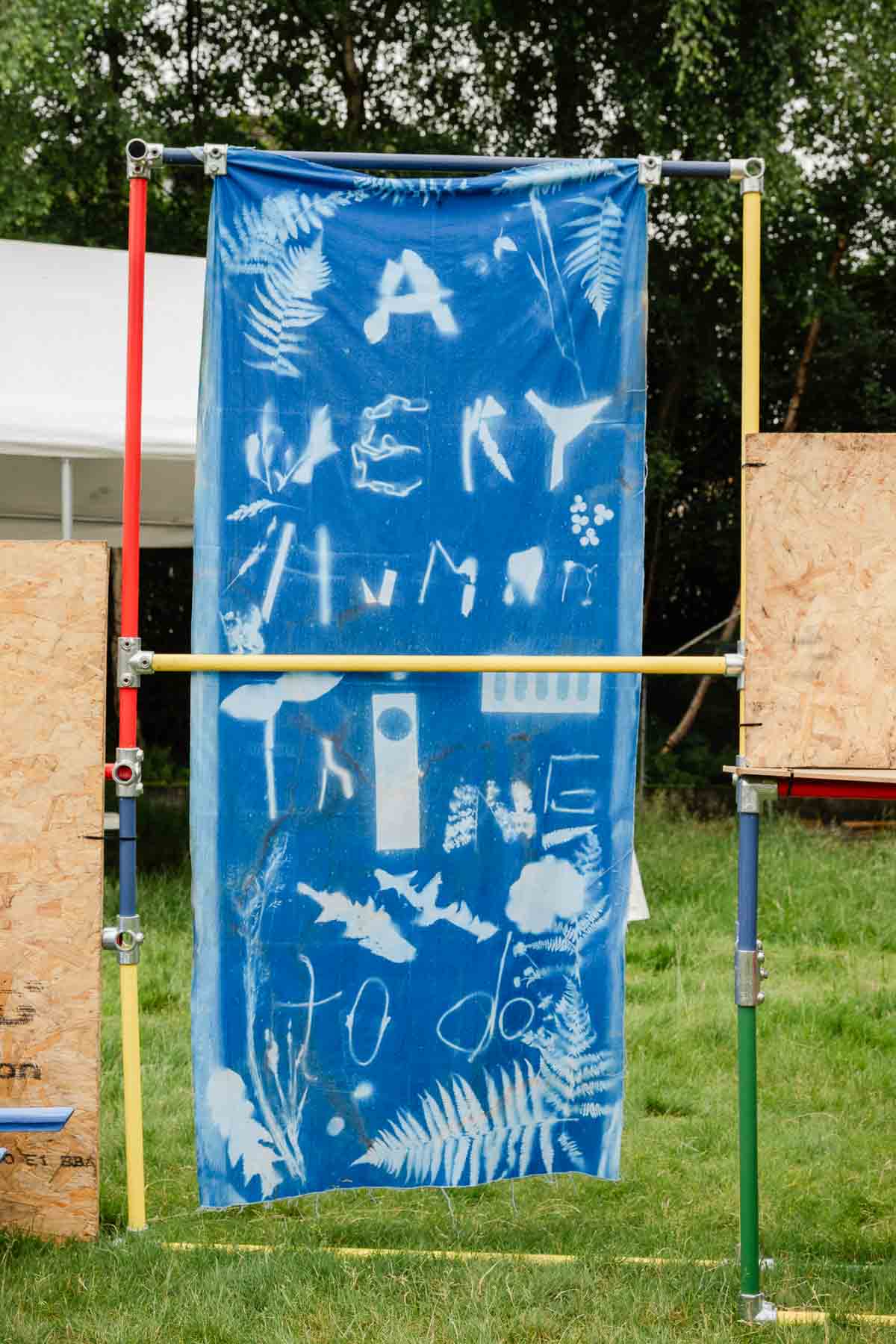 A blue cyanotype banner reading "A Very Human Thing To Do" with white silhouettes of ferns and leaves, hung on a scaffolding-pole frame in a grassy field.