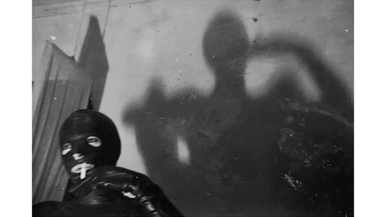 Black-and-white photography. On the bottom left is a person smoking and wearing a latex suit, their shadow casts on to the wall in the background. 