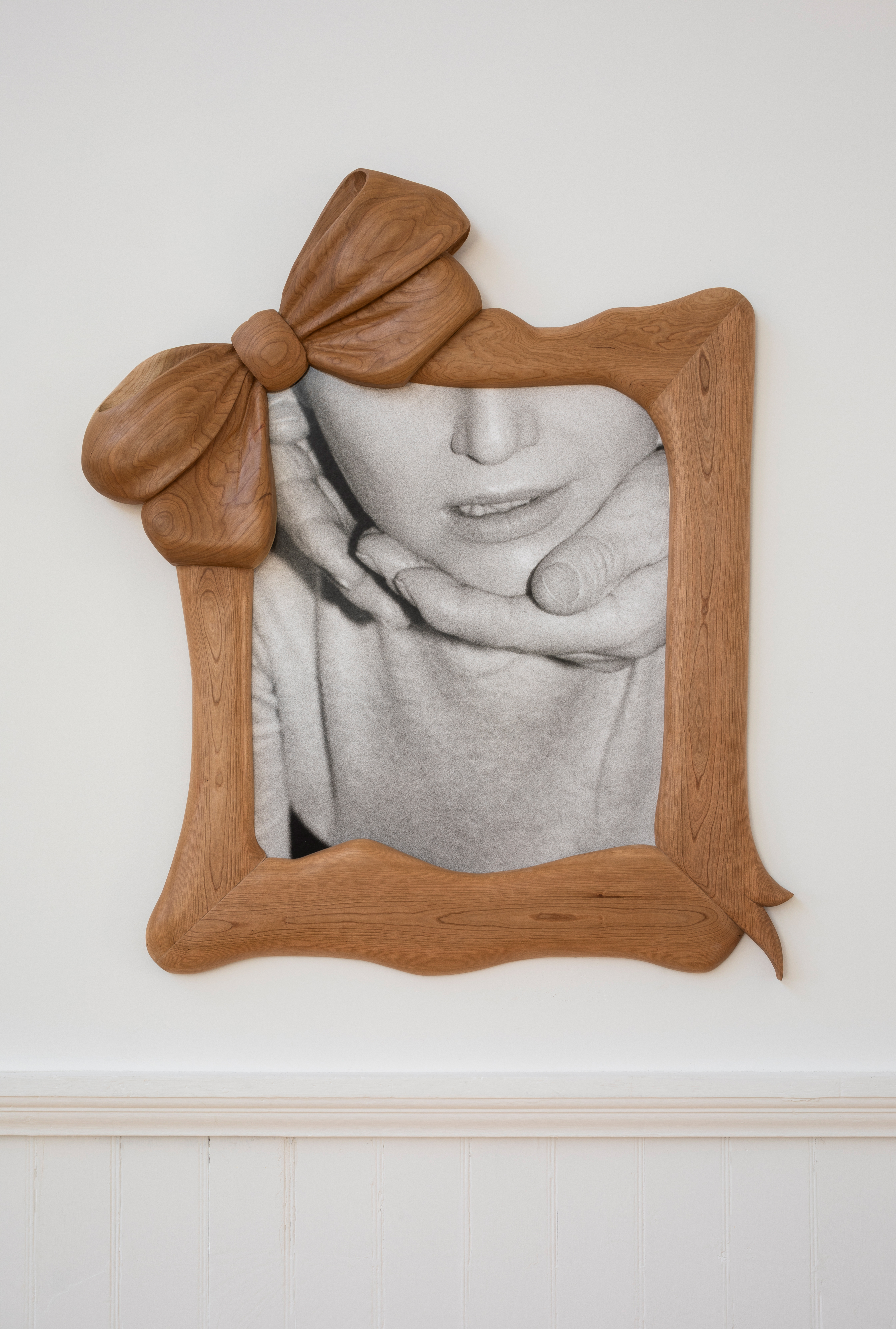 A carved wooden bow, styled like a decorative frame, sits atop a black and white photograph showing a hand cradling someone's chin.