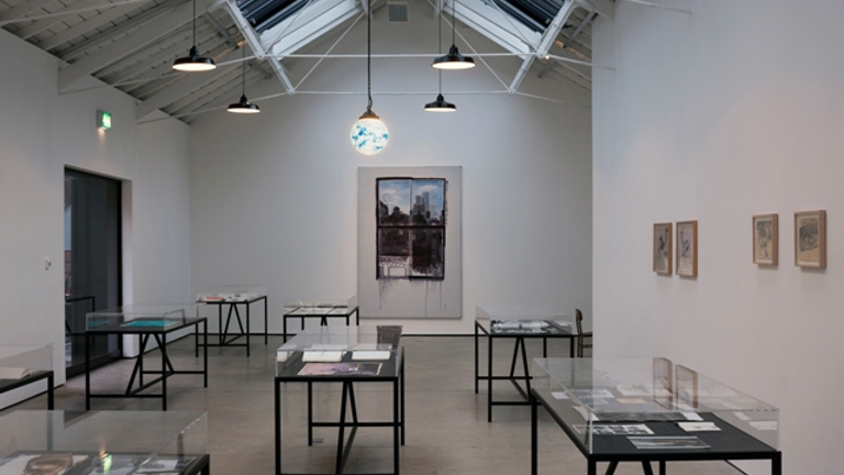 gallery space with multiple black metal tables encased by glass tops. they contain notebooks. on the back wall there is a large canvas with a painting of a view from a window. on the right wall there are four framed drawings.