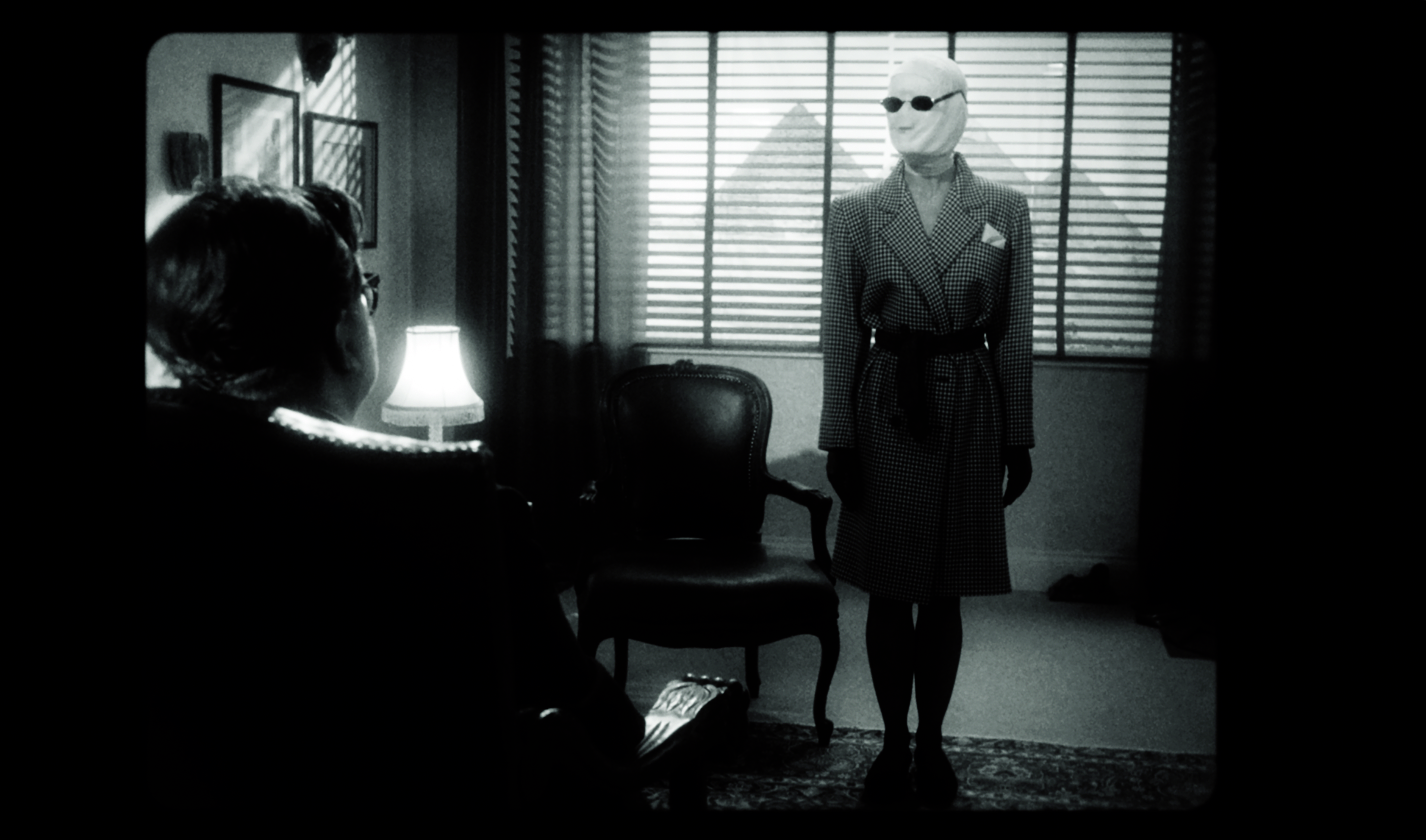 Black-and-white film still of a psychoanalytic session. In the foreground, a figure sits in shadow with their back to the camera, facing another figure standing beside benetian-blinded windows. The standing figure wears a tailored 1940s checkered suit, dark sunglasses, and has their head fully wrapped in bandaded. The room it lit by a table lamp, with framed artworks on the walls, evoking film noir aesthetics and the clincal atmosphere of mid-century psychoanalysis