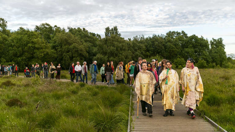 Three people wearing off white cloaks lead a group of people through a boggy peatland on a wooden walkway.